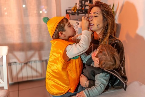 Two young brothers in halloween costumes playfully applying makeup on their mother's face in a