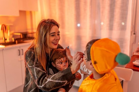 Happy family preparing for halloween party, mother applying makeup on children's faces, creating