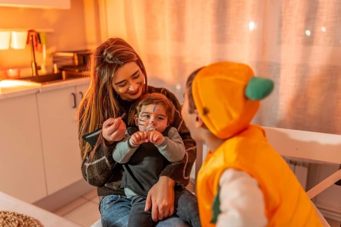 Mother applying halloween makeup on her toddler son dressed up as a cat while his brother dressed