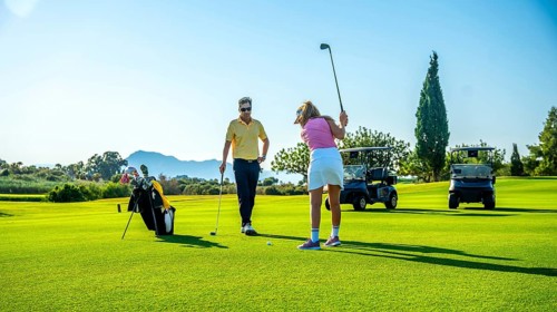 Golf instructor coaching female golfer on driving range, with golf carts in background on sunny day