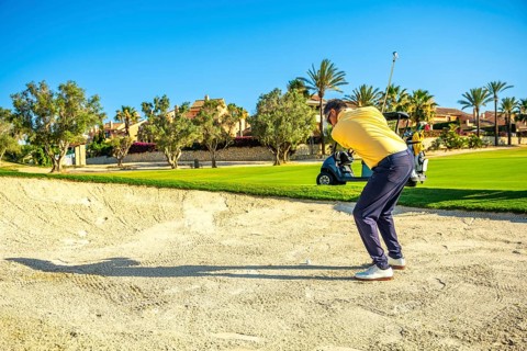 Golfer concentrating on hitting ball out of sand trap on a beautiful sunny day, with golf cart in