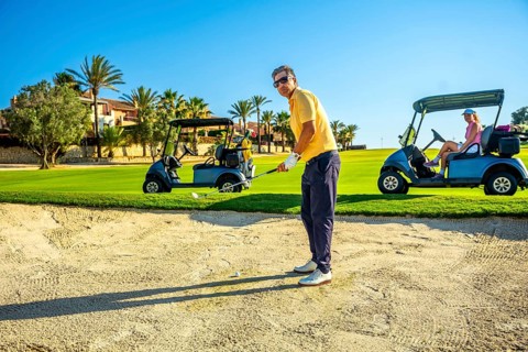 Golfer focuses on a bunker shot while others relax in golf carts on a bright, sunny day at a lush