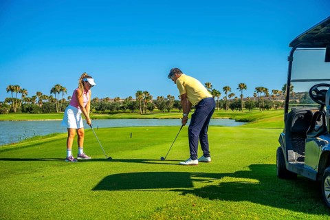 Father and daughter duo playing golf on a sunny day, surrounded by palm trees and a clear blue sky,
