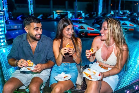 Three friends are enjoying sandwiches while sitting on a bumper cars platform, illuminated by