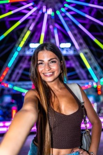 Happy young woman taking a selfie with a colorful ferris wheel in the background at an amusement