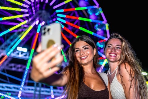 Two happy women taking a selfie with smartphone in front of illuminated ferris wheel at an