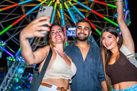 Three happy friends taking a selfie with smartphone in front of a colorful ferris wheel at an