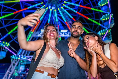Three friends taking a selfie with a smartphone in front of a brightly lit ferris wheel at an