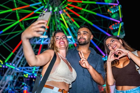 Three friends taking a selfie with smartphone in front of a colorful ferris wheel, enjoying