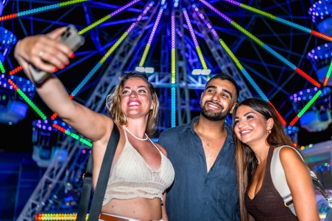 Three happy friends taking a selfie with smartphone in front of a colorful ferris wheel at an