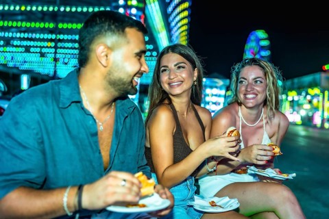 Happy friends sharing delicious hot dogs while enjoying the vibrant atmosphere of an amusement park