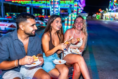 Three friends are sitting and enjoying hot dogs together at an amusement park, illuminated by