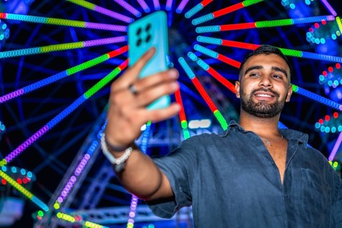 Tourist taking selfie with smartphone in front of illuminated ferris wheel at amusement park,