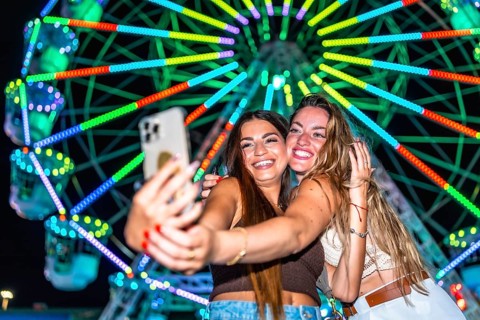 Two young women taking a selfie with a smartphone in front of a brightly lit ferris wheel at an