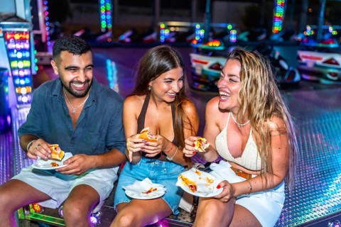 Three friends are sitting and laughing while eating hot dogs at an amusement park's bumper cars