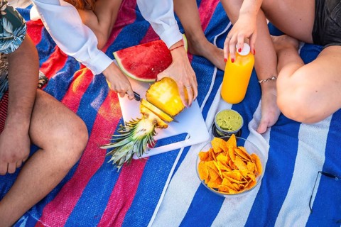 Friends are having a picnic on a striped towel, cutting pineapple, with watermelon, nachos,