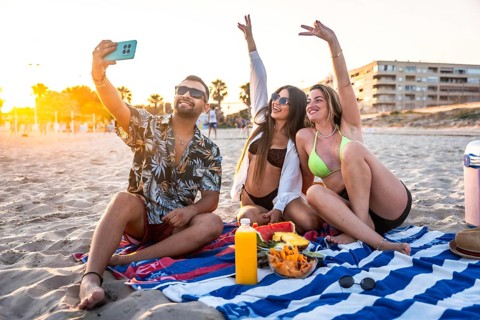 Three friends are sitting on a beach towel, taking a selfie while enjoying snacks and drinks during