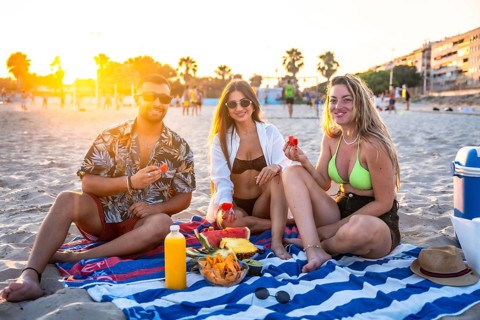 Three friends enjoying a picnic on the beach at sunset, eating watermelon and other fresh fruit