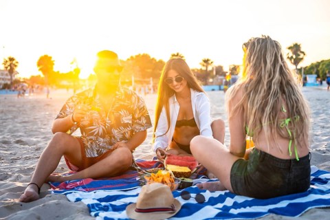 Group of friends enjoying watermelon and pineapple during a sunset picnic on a beach towel,