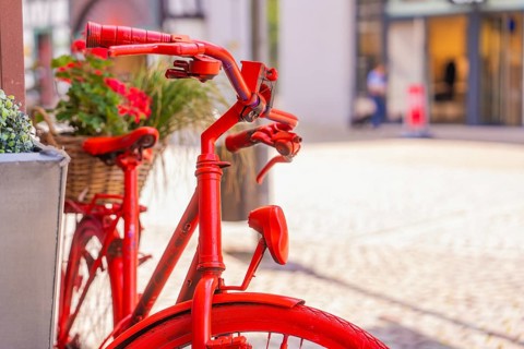 Close-up of a red bicycle handlebar with flower baskets in a lively city scene, small town of Perle