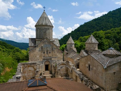 Historic stone monastery surrounded by green forests under a blue sky with clouds, aerial view,