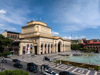 An impressive building dominates the square, flanked by a fountain and urban life, aerial view,