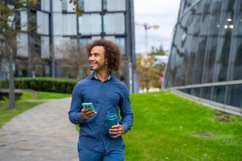 Smiling young Mixed-race man with curly afro hair walking on a path in a modern city park, holding