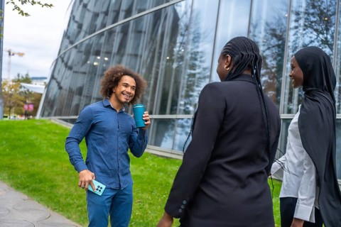 Diverse colleagues are enjoying a casual coffee break outdoors, talking and smiling against a