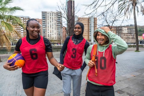Three diverse friends, a young woman holding a basketball and two people wearing red bibs, walking