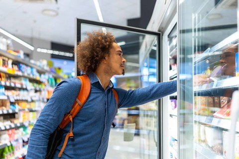 Young man shopping in a grocery store, reaching for frozen items in a refrigerated display,