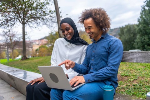 Diverse young couple sitting on a park bench, smiling and pointing at a laptop screen while sharing