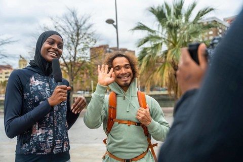 Smiling female journalist wearing a hijab holding a microphone, interviewing a happy man waving on