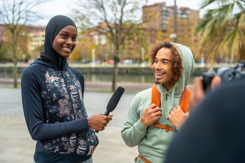 Female journalist wearing a hijab holding a microphone, interviewing a cheerful young man with