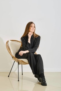 A young woman wears a oversized black outfit while seated comfortably in a modern chair. Her