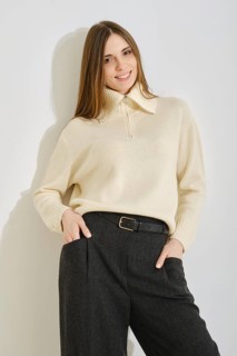 A woman stands by the bright wall with a warm smile, wearing a stylish cream sweater and loose dark