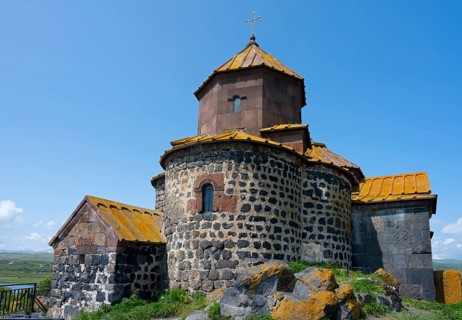 Historic stone church with a round tower in a rural setting under blue sky, Hajravank Monastery,