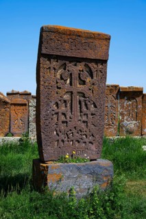 A detailed Armenian crossstone with engravings in front of several others, surrounded by grass