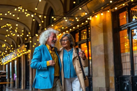 Senior couple walking arm in arm under string lights, happily talking while enjoying an evening out