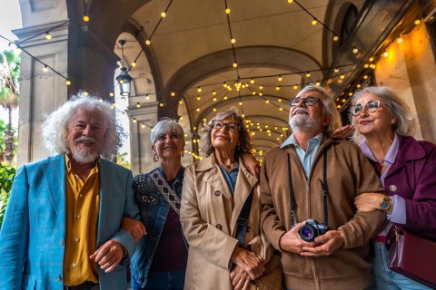 Senior friends strolling beneath a decorated arcade, smiling and looking up as they enjoy a sunny