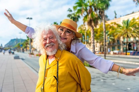 Senior couple laughing as husband gives wife a piggyback through sunlit city streets lined with