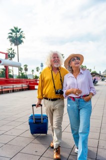Senior couple walking on a city promenade, smiling and looking up, carrying a suitcase and a
