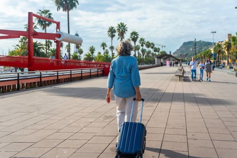 Senior woman walking with a suitcase along a sunny boardwalk lined with palm trees and a