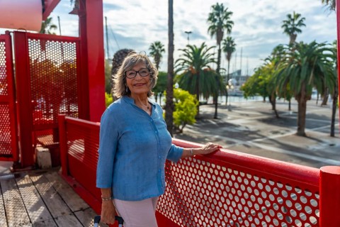 Senior woman standing on red bridge at a sunny harbor, smiling confidently in glasses while