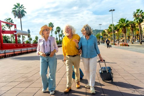 Three cheerful senior tourists walking together on a promenade with palm trees, carrying luggage