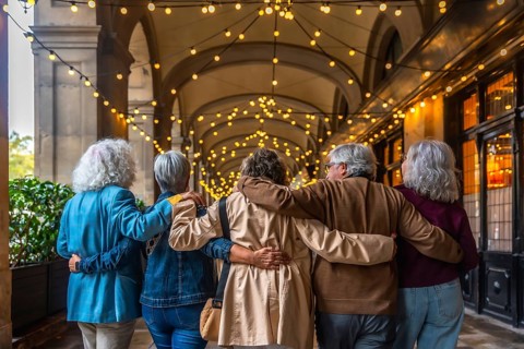 Group of diverse senior friends walking together embraced along an illuminated city archway at