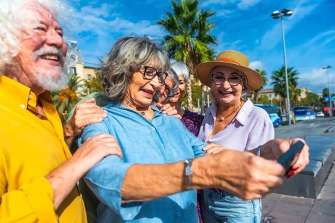 Group of smiling seniors enjoying a vacation, standing outdoors together and using a smartphone to