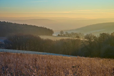 Frozen meadow in morning light with a foggy valley and rolling hills in the background, Seckmauern,