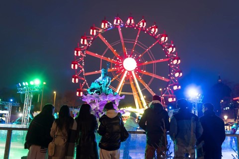 Several people watch ice skaters in front of the Neptune Fountain and a Ferris wheel at the Berlin