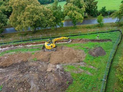 Excavator on a construction site with earthworks next to a road and trees, Bau PV