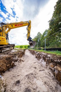 A yellow excavator digs a deep ditch surrounded by nature and a cloudy sky, Bau PV